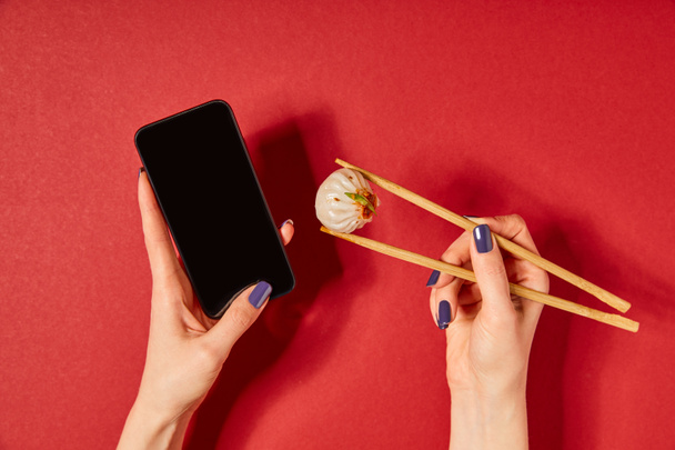 Draufsicht der Frau mit Essstäbchen mit gedämpftem Brötchen und Smartphone mit leerem Bildschirm auf Rot - Foto, Bild