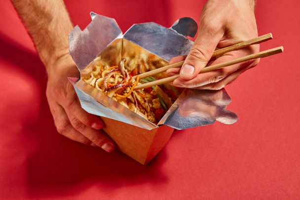 cropped view of man holding chopsticks near tasty noodles on red - Foto, afbeelding