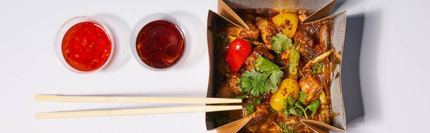 panoramic shot of sauces and chopsticks near takeaway box with prepared pork and vegetables on white  - Foto, immagini