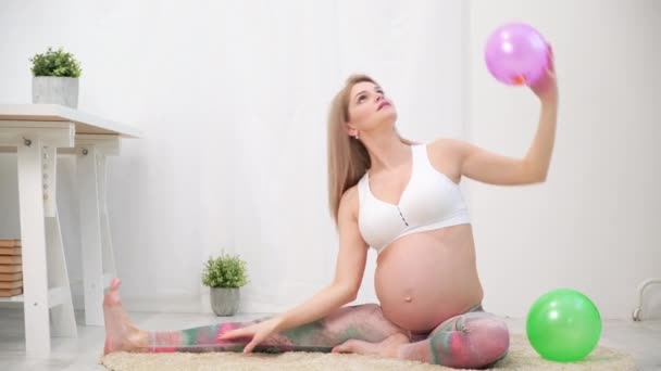 A young beautiful pregnant Caucasian woman sits on the soft carpet of the house. He is engaged in fitness and doing exercises. Sportswear. - Séquence, vidéo