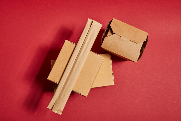Ansicht von Essstäbchen in Papierverpackungen in der Nähe von Kartons zum Mitnehmen mit traditionellen chinesischen Lebensmitteln auf Rot - Foto, Bild