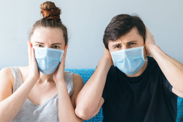 Married couple in blue medical face masks sitting on the couch, holding their heads with their hands. Man and woman in shock, family of two, stay at home during quarantine - Fotografie, Obrázek