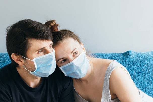 Getrouwd echtpaar in blauw medisch gezicht maskers zitten op de bank, knuffelen en helaas kijken in de verte. Man en vrouw, familie van twee, thuis blijven tijdens quarantaine - Foto, afbeelding