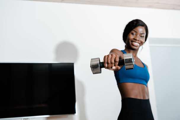African american sportswoman holding dumbbell with outstretched hand, smiling and looking at camera in living room - Photo, Image