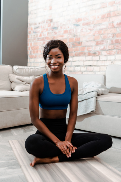 Afrikai amerikai sportos nő lótuszban pózol mosolyogva és a kamerát nézi jóga szőnyegen a nappaliban - Fotó, kép