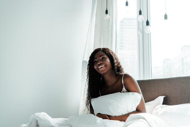 Donna afroamericana che tiene cuscino e sorride sul letto in camera da letto
 - Foto, immagini