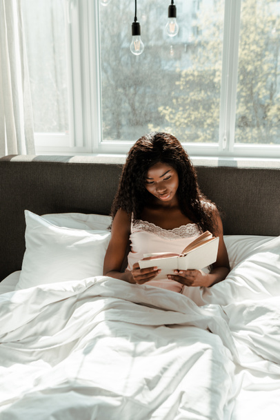 African american woman reading book on bed in bedroom - Fotó, kép
