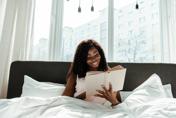 African american woman reading book and smiling on bed in bedroom - Foto, imagen