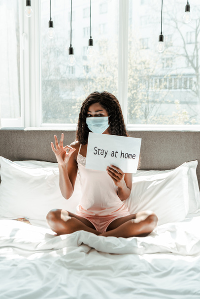Αφροαμερικανή γυναίκα σε ιατρική μάσκα με σταυρωμένα πόδια δείχνει εντάξει σημάδι και πλακάτ με διαμονή στο σπίτι επιγραφή στο κρεβάτι στο υπνοδωμάτιο - Φωτογραφία, εικόνα