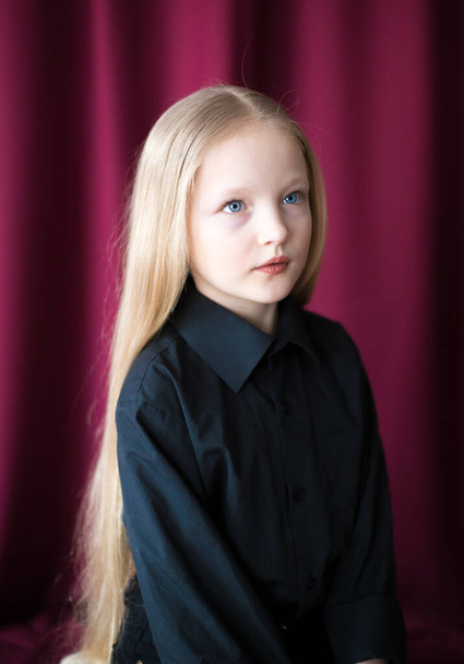 Cute little girl with long blond hair in a black shirt and jeans on a red background in the studio - Fotografie, Obrázek