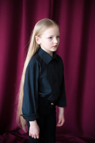 Cute little girl with long blond hair in a black shirt and jeans on a red background in the studio - Foto, imagen