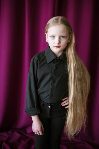 Cute little girl with long blond hair in a black shirt and jeans on a red background in the studio - Fotó, kép