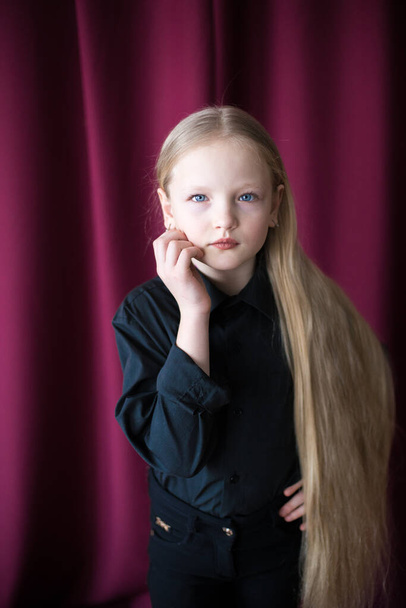 Cute little girl with long blond hair in a black shirt and jeans on a red background in the studio - Valokuva, kuva
