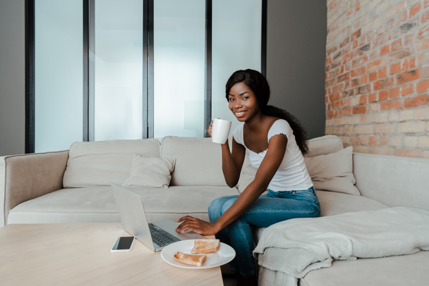 African american freelancer with cup of tea smiling and using laptop at coffee table with smartphone and plate with toasts in living room - Foto, Bild
