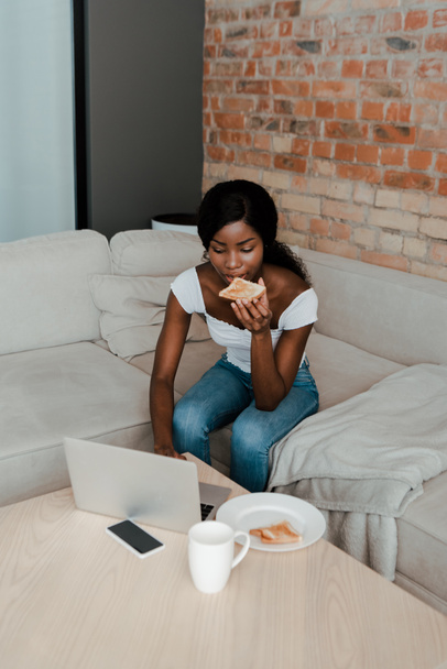 freelancer afroamericano comiendo tostadas y usando portátil en la mesa de café con teléfono inteligente, plato y taza de té en la sala de estar
 - Foto, imagen