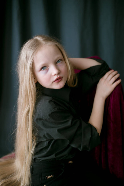 Linda niña con el pelo largo y rubio en una camisa negra y jeans sobre un fondo negro en el estudio
 - Foto, imagen