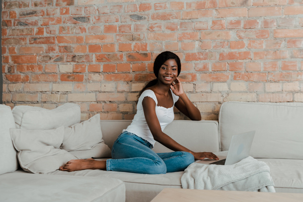 African american freelancer using laptop, smiling and looking at camera on sofa in living room  - Foto, imagen