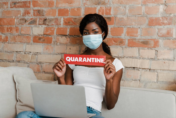 Afrykański freelancer amerykański w masce medycznej z laptopem pokazujący kartę z napisem kwarantanny i patrząc na kamerę na kanapie - Zdjęcie, obraz
