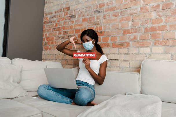 Afrikai amerikai szabadúszó orvosi maszk laptop mutatja kártya karantén betűkkel és ellenszenv jel kanapén - Fotó, kép