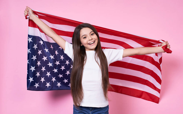 Funny stylish young emotional girl, long-haired brunette, smiles broadly and holds joyfully American flag, Independence Day, July 4, victory,peace,USA, in a white base T-shirt on a pink background. - Foto, Imagen