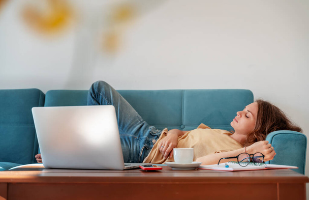 Charming beautiful young girl in glasses with curly hair fell asleep on a blue sofa at home in front of a laptop, notepad and a cup of coffee, remote work and education. Fatigue, Deadline - Photo, image