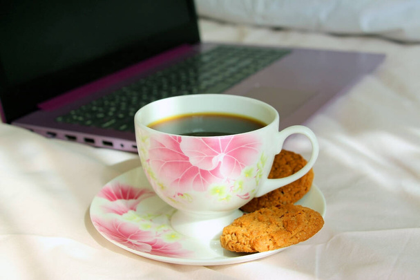 A cup of tea with cookies on a pink bed with a purple laptop. Remote work at home. - Photo, Image