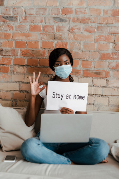 African american freelancer in medical mask with laptop showing okay sign and placard with stay at home lettering on sofa in living room - Фото, изображение