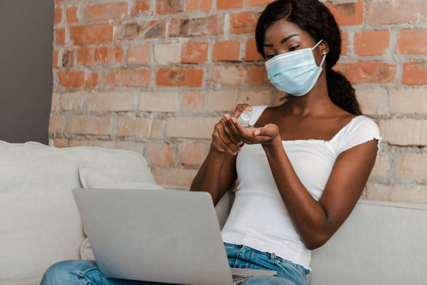 African american freelancer in medical mask with laptop using hand sanitizer on sofa in living room - Foto, Imagen