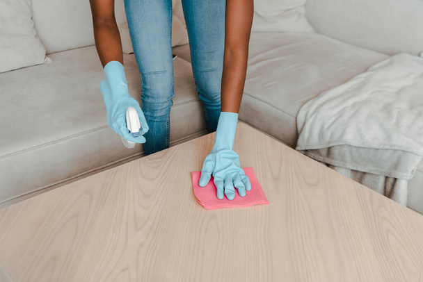 Cropped view of african american woman in rubber gloves cleaning coffee table with detergent and rag in living room - Фото, зображення