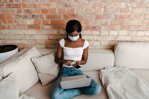Hochblickaufnahme eines afrikanisch-amerikanischen Freiberuflers in medizinischer Maske mit desinfizierendem Laptop-Smartphone mit Händedesinfektionsmittel auf dem Sofa  - Foto, Bild