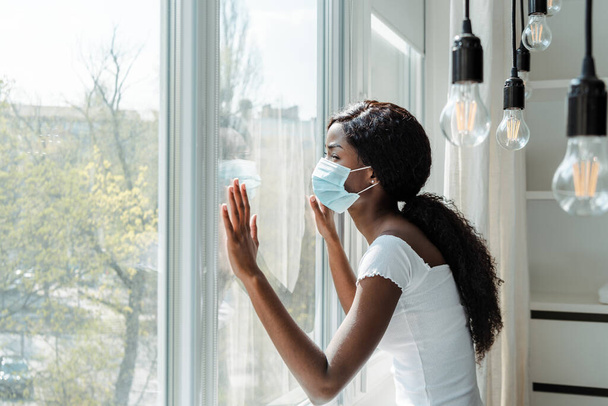 African american woman in medical mask touching windows in living room - Photo, Image