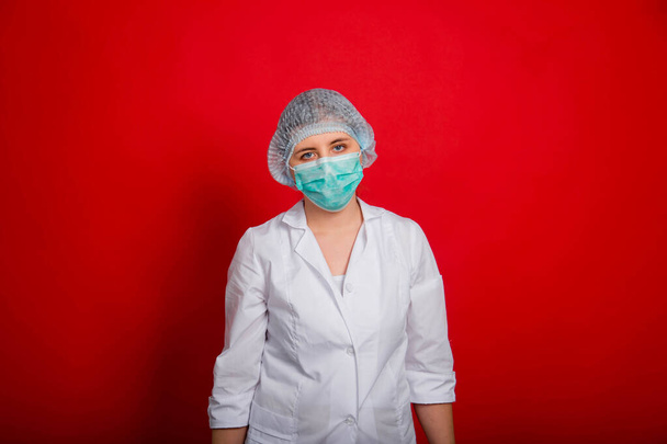 Уставшая женщина-врач в белом халате, маске и медицинской шапке стоит на красном фоне
. - Фото, изображение