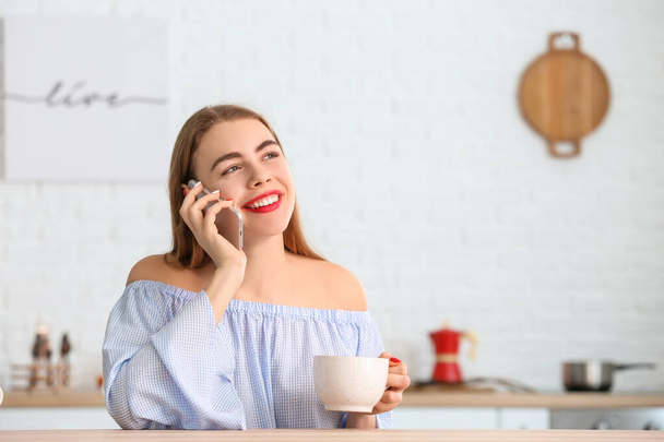 Piękna młoda kobieta pije herbatę i rozmawia przez telefon w kuchni - Zdjęcie, obraz