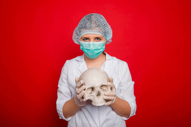 Una doctora en ropa médica sostiene un cráneo en sus manos mientras está de pie sobre un fondo rojo
. - Foto, Imagen