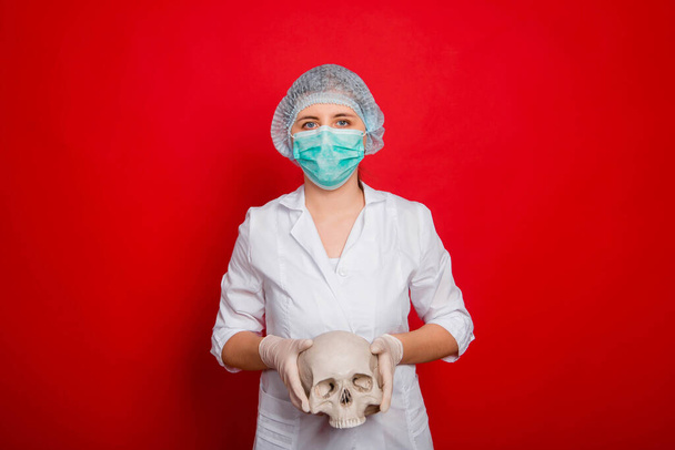 A woman doctor in medical clothes holds a skull in his hands while standing against a red background. - Photo, Image