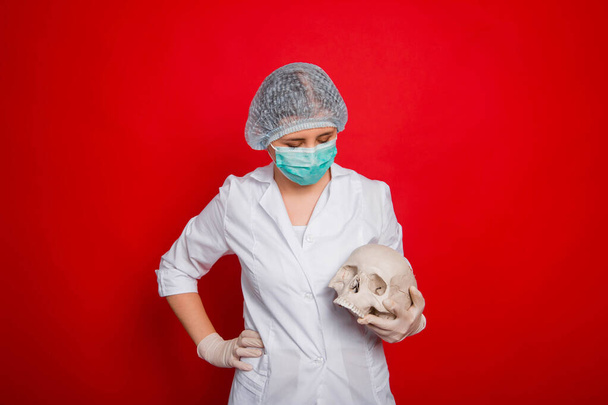 Μια γυναίκα γιατρός με ιατρικά ρούχα κρατά ένα κρανίο στα χέρια του ενώ στέκεται πάνω σε ένα κόκκινο φόντο. - Φωτογραφία, εικόνα