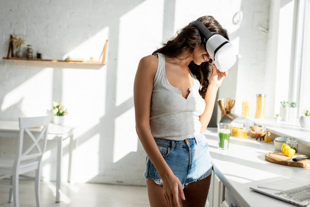 Foco seletivo de mulher sorridente em fone de ouvido realidade virtual em pé perto de laptop na cozinha
  - Foto, Imagem