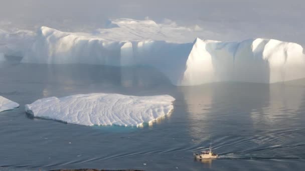 Een kleine boot onder ijsbergen. Zeilboot cruisen tussen drijvende ijsbergen in Disko Bay Glacier tijdens Midnight Sun Ilulissat, Groenland. Studeren van een fenomeen van de opwarming van de aarde ICES en ijsbergen - Video