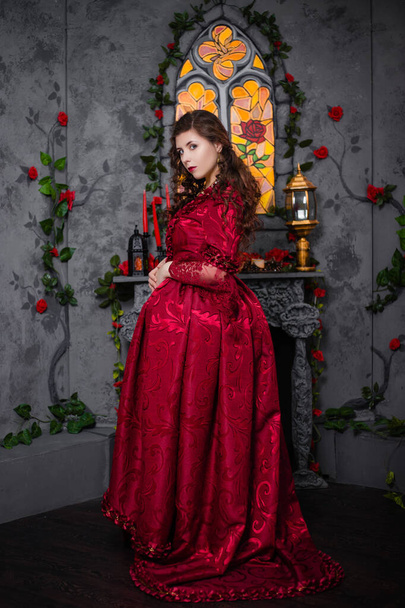 Beautiful girl in a magnificent, red rococo dress. Against the background of the fireplace, window, and flowers. - Foto, immagini