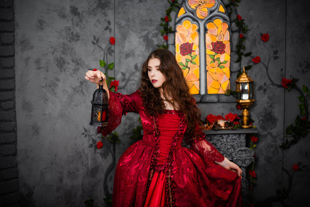 A beautiful girl in a magnificent red dress of the Rococo era stands against a fireplace, a window and flowers with a lamp with candles in her hands. - Fotoğraf, Görsel