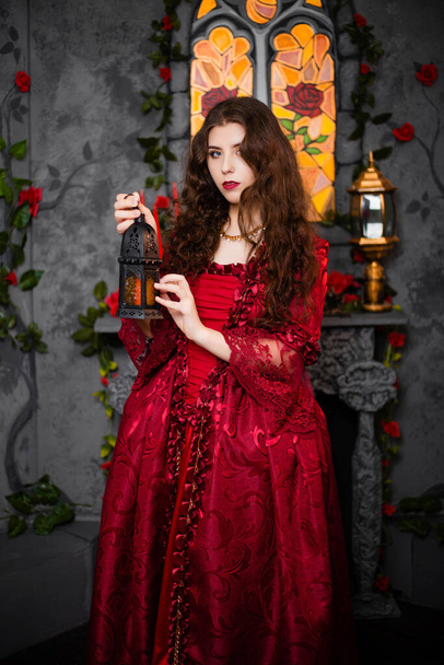 A beautiful girl in a magnificent red dress of the Rococo era stands against a fireplace, a window and flowers with a lamp with candles in her hands. - Foto, Imagem