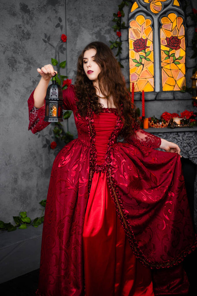 A beautiful girl in a magnificent red dress of the Rococo era stands against a fireplace, a window and flowers with a lamp with candles in her hands. - Fotó, kép