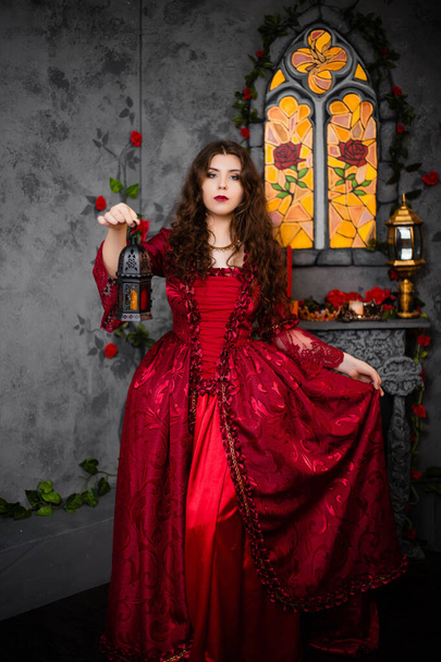 A beautiful girl in a magnificent red dress of the Rococo era stands against a fireplace, a window and flowers with a lamp with candles in her hands. - Фото, изображение
