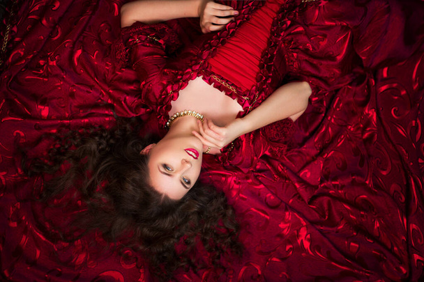 A beautiful young girl lies on a magnificent red dress of the Rococo era. - Fotografie, Obrázek