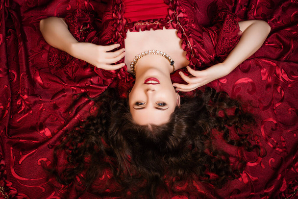 A beautiful young girl lies on a magnificent red dress of the Rococo era. - Photo, Image