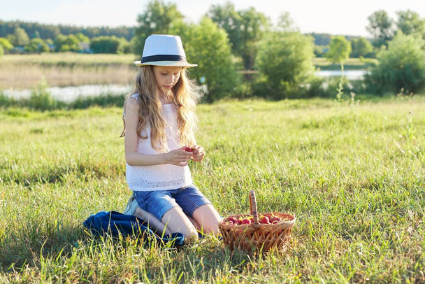 Outdoor summer portrait of little girl with basket strawberries, straw hat. Nature background, rural landscape, green meadow, country style, child sitting on the grass eating berries - Photo, image