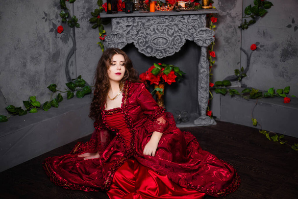 A beautiful girl in a magnificent red dress of the Rococo era sits on the floor near a fireplace and flowers. - Fotografie, Obrázek
