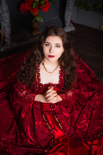 A beautiful girl in a magnificent red dress of the Rococo era sits on the floor near a fireplace and flowers. - Фото, изображение