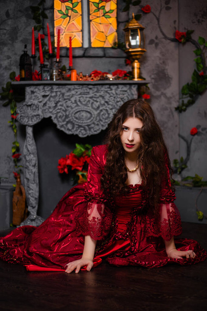 A beautiful girl in a magnificent red dress of the Rococo era sits on the floor near a fireplace and flowers. - Valokuva, kuva