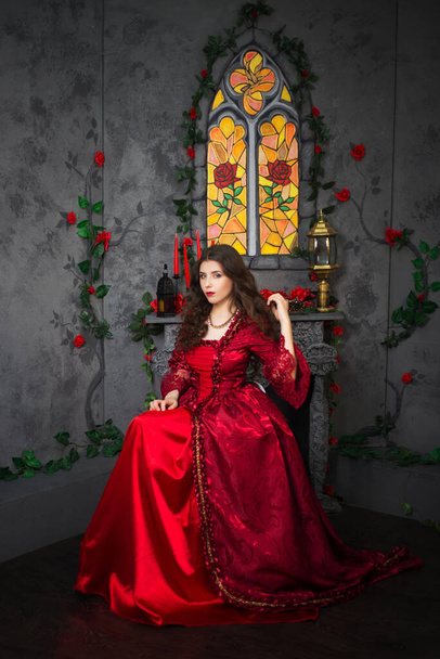 A beautiful girl in a magnificent red dress of the Rococo era sits on a chair near the fireplace, window and flowers. - Foto, Imagen